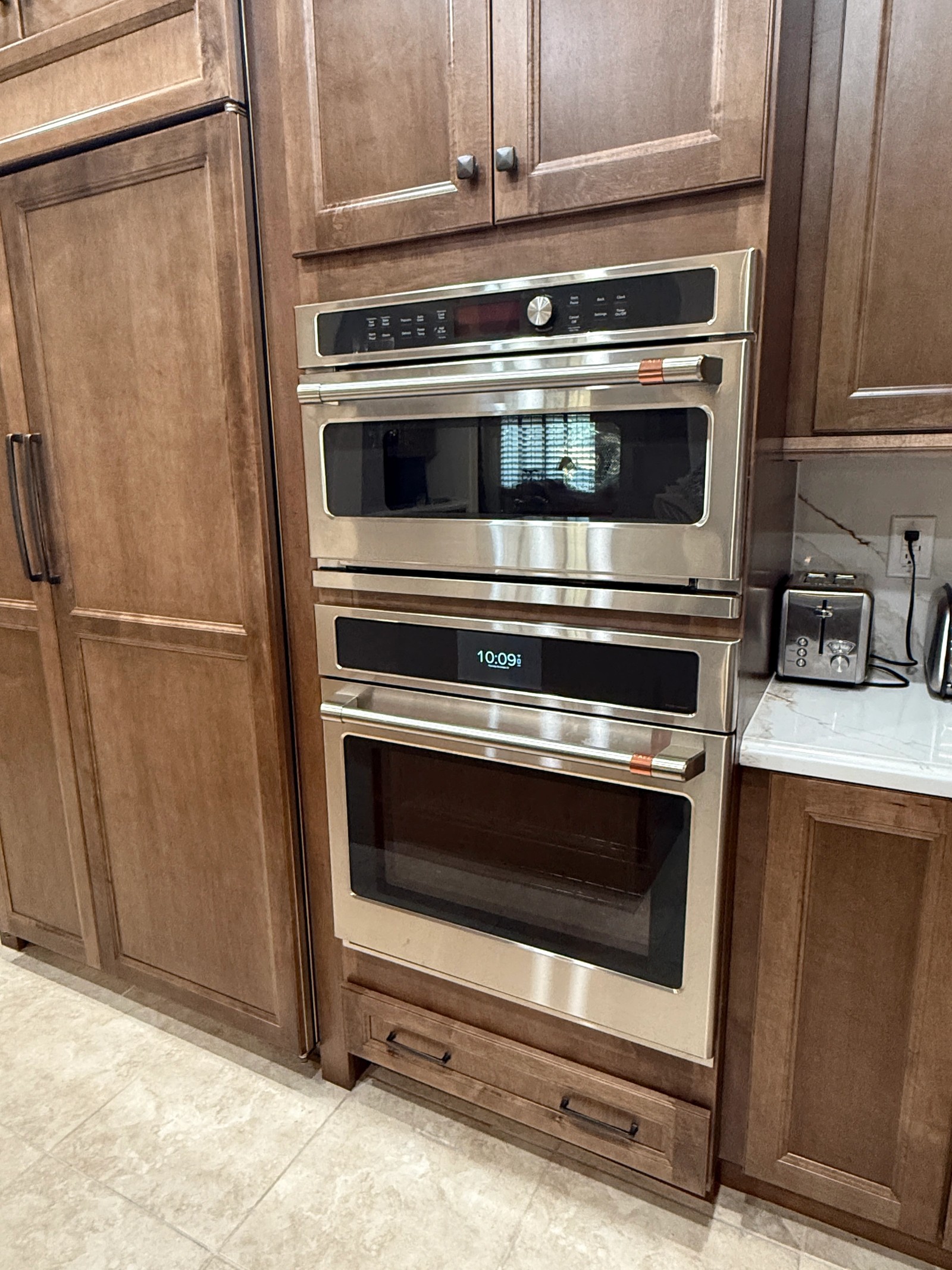 Stacked stainless wall ovens set into a tall stained maple cabinet run beside a panelled refrigerator